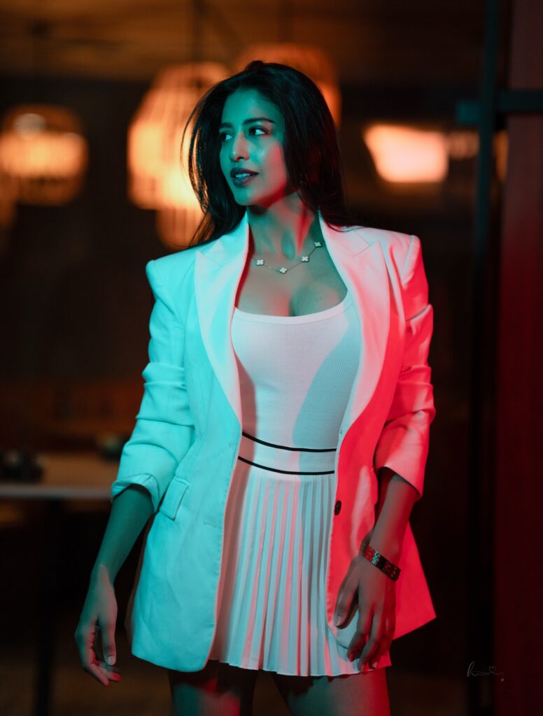Daksha Nagarkar in a sexy white blazer and pleated mini skirt, glowing under neon lights, showcasing her stunning legs and figure