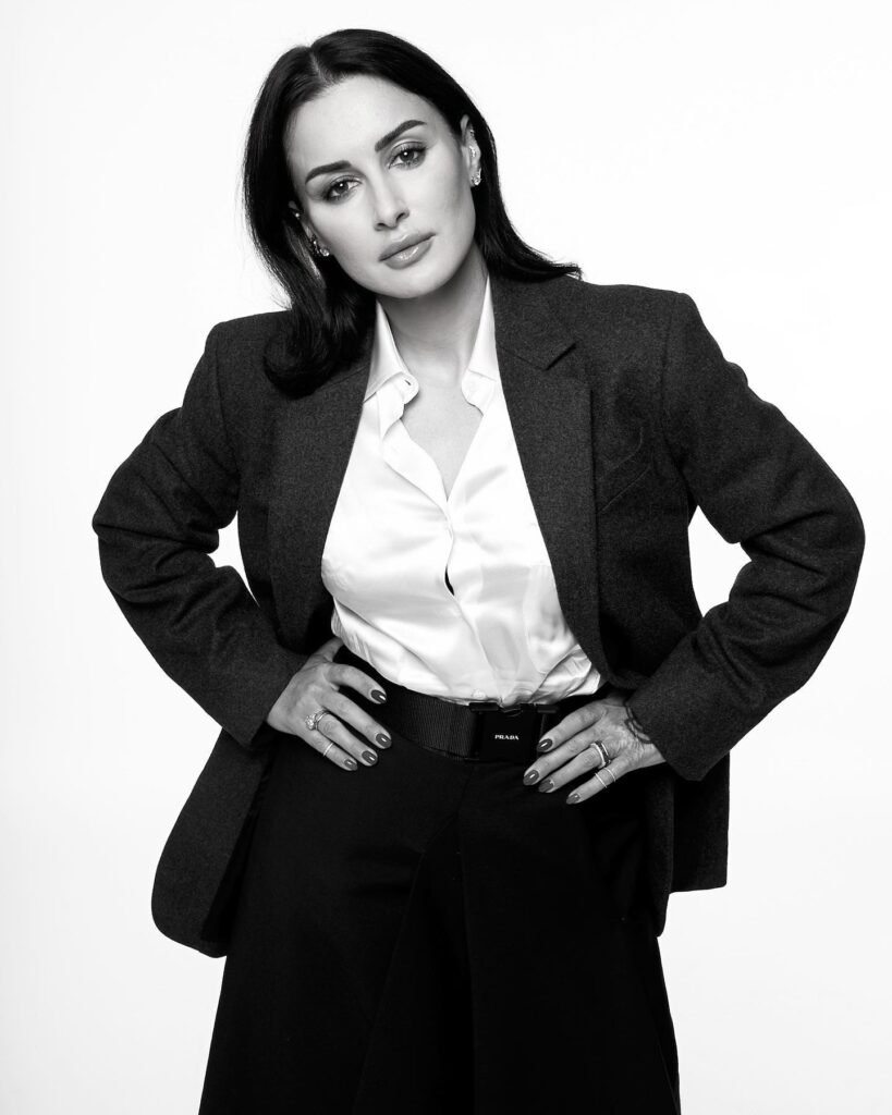 The black-and-white photo shows Tina Kandelaki in a tailored blazer, silky blouse, high-waisted skirt with a belt, and high heels. Accessorized with rings, she stands confidently, hands adjusting the blazer, exuding elegance against a plain background. Let me know if you need further assistance!