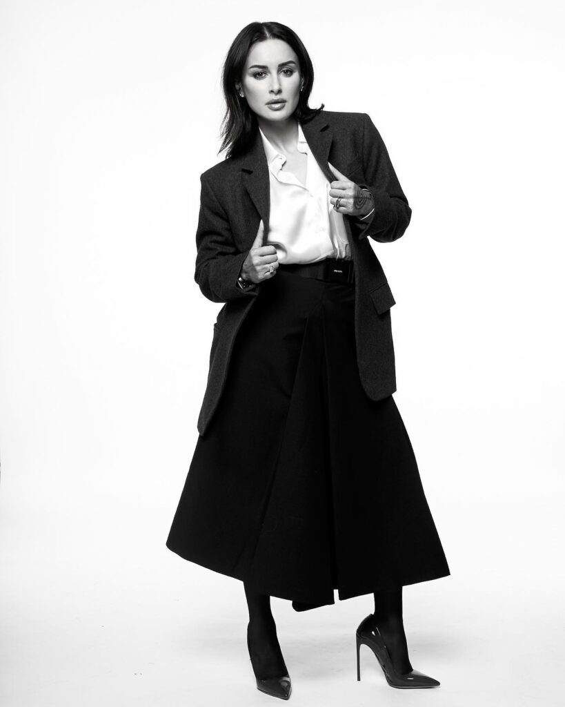 The black-and-white photo shows Tina Kandelaki in a tailored blazer, silky blouse, high-waisted skirt with a belt, and high heels. Accessorized with rings, she stands confidently, hands adjusting the blazer, exuding elegance against a plain background. Let me know if you need further assistance!